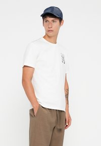 White cotton t-shirt with round neckline and short sleeves, featuring a small black graphic pocket print. Paired with brown textured pants.