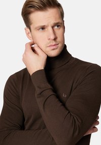 Brązowy, prążkowany sweter z golfem z dopasowanym krojem. Subtelny detal z logo na klatce piersiowej. Tekstura wydaje się miękka i ciepła.