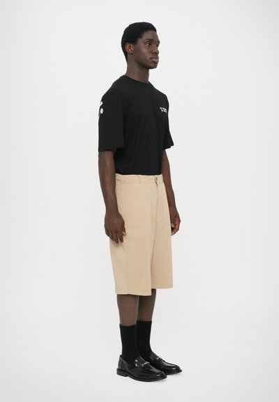 T-shirt noir à manches courtes, shorts beige jusqu'aux genoux, chaussettes de costume noires, mocassins noirs brillants. Design minimaliste, sans motifs ni graphiques visibles.