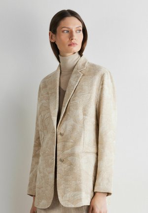 Frau trägt ein beige gemustertes Blazer über einem beigen Rollkragenpullover und blickt seitlich vor einem schlichten weißen Hintergrund.