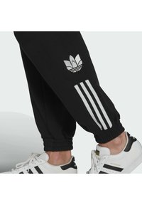 Underben som bär svarta Adidas-byxor med vita tre ränder och logotyp, tillsammans med vita Adidas Superstar-sneakers med svarta ränder.