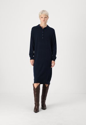 Navyblaues Langarmkleid mit Polo-Kragen, Knopfleiste, seitlichen Schlitzen und gerippten Bündchen, kombiniert mit braunen Cowboyschnürstiefeln mit geprägten Details.