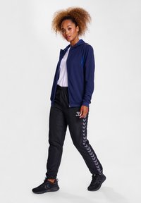 Marineblauwe zip-up jas met blauwe accenten, wit T-shirt, zwarte trainingsbroek met visgraatpatroon en zwarte sportschoenen.