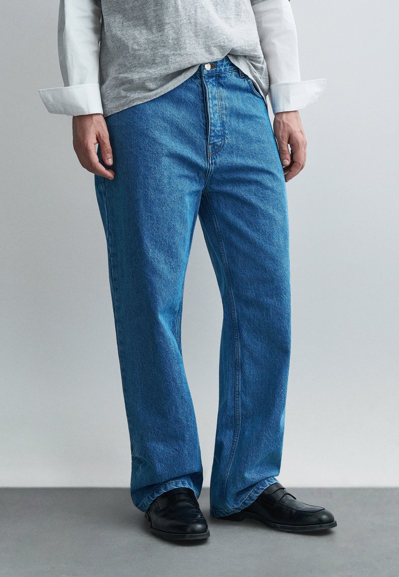 Sinised teksad sirge säärega, millel on nööpidega läbilöök ja kergelt pleekinud toon. Kandmisel hall T-särk ja mustad kingad.