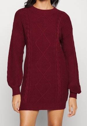 Femme portant une robe en tricot bordeaux à manches longues avec des motifs en câble et en diamant, ainsi que des poignets et un ourlet côtelés.