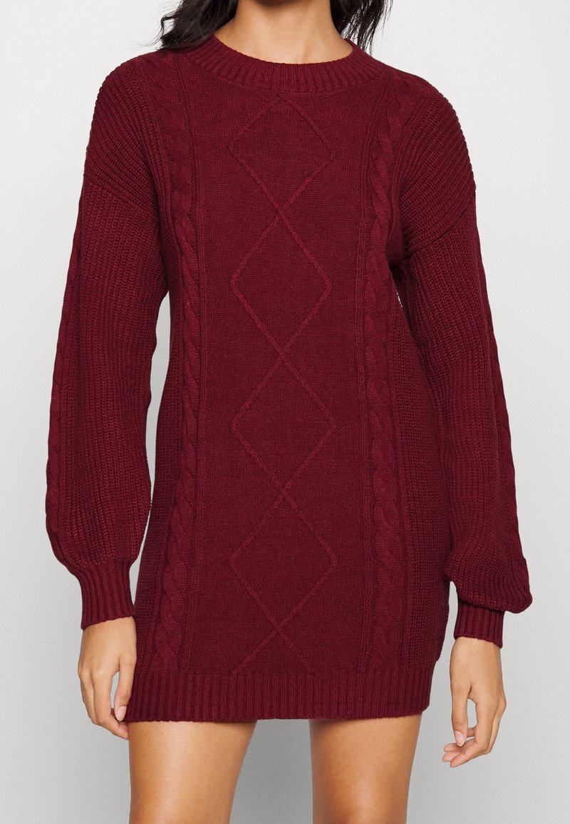 Donna che indossa un vestito di maglione a maniche lunghe di colore bordeaux con motivi a treccia e diamante e polsini e orlo a coste.