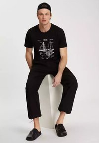 Czarny t-shirt z białą grafiką łódki żaglowej, w zestawieniu z czarnymi spodniami i czarnymi sandałami, siedzi na białym trójwymiarowym bloku.