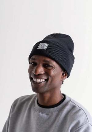 Sort ribbet hue med et mærketikette; blød tekstur. Bæres med en grå sweatshirt med rund halsudskæring og kontrastfarvede manchetter.