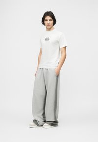 Νεαρός στέκεται με τα χέρια στις τσέπες, φοράει λευκό T-shirt με λογότυπο, υπερμεγέθη, ανοιχτόγριες φόρμες και λευκά αθλητικά παπούτσια σε απλό φόντο.