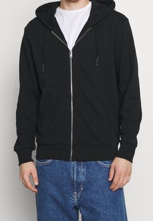 Mand iført en sort hættetrøje med lynlås, en hvid t-shirt under og blå jeans, stående mod en ensfarvet baggrund.