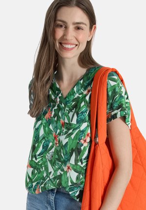 Uśmiechnięta kobieta z długimi brązowymi włosami, ubrana w zieloną koszulę w kwiaty i niebieskie dżinsy, niesie dużą pomarańczową pikowaną torbę na ramieniu.