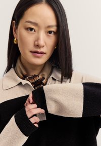 Junge Frau, die den Ärmel eines schwarz-beige gestreiften Pullovers anpasst, trägt einen braunen gemusterten Halstuch und goldene Creolen, neutraler Hintergrund.