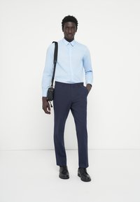 Svetlo modra srajca z gumbi, mornarske hlače in črne oblečne čevlje. Model nosi črno torbico preko ramena, stoji ob enaki ozadju.