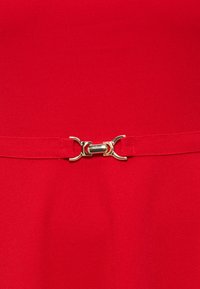 Κοντινή εικόνα κόκκινης υφασμάτινης ζώνης με κούμπωμα από χρυσαφί μέταλλο στερεωμένο στο κέντρο.