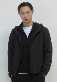 Giacca nera con zip e cappuccio, caratterizzata da una texture liscia e una silhouette aderente. Indossata sopra un top nero stratificato con una camicia bianca.