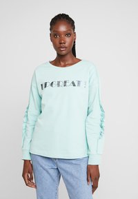 Sweatshirt verde menta com impressão em material refletivo "UPGREAT!". Apresenta mangas franzidas e decote redondo, combinado com jeans de denim claro.