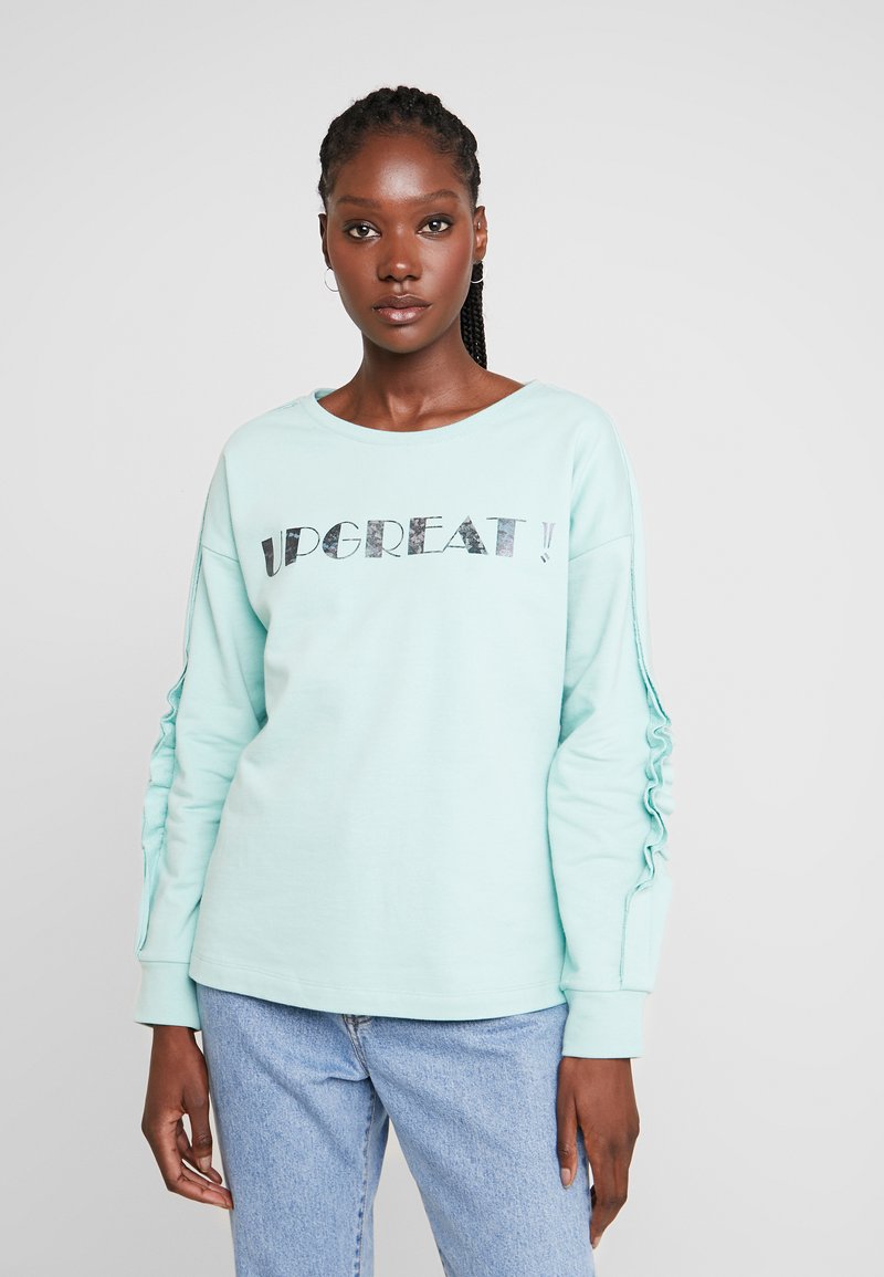 Sweatshirt verde menta com impressão em material refletivo "UPGREAT!". Apresenta mangas franzidas e decote redondo, combinado com jeans de denim claro.