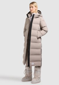 Vrouw in een lange beige pufferjas met een capuchon, met haar handen in de zakken, beige enkellaarsjes met veters voor de winter, staand tegen een witte achtergrond.