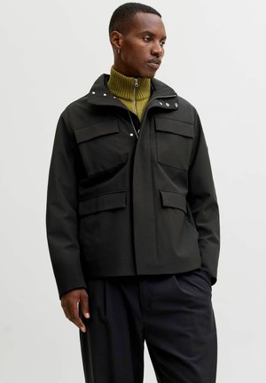 Svart vattentät jacka med hög krage, framdragkedja, tryckknappar och två stora bröstfickor. Bärs över en grön ribbad polotröja.