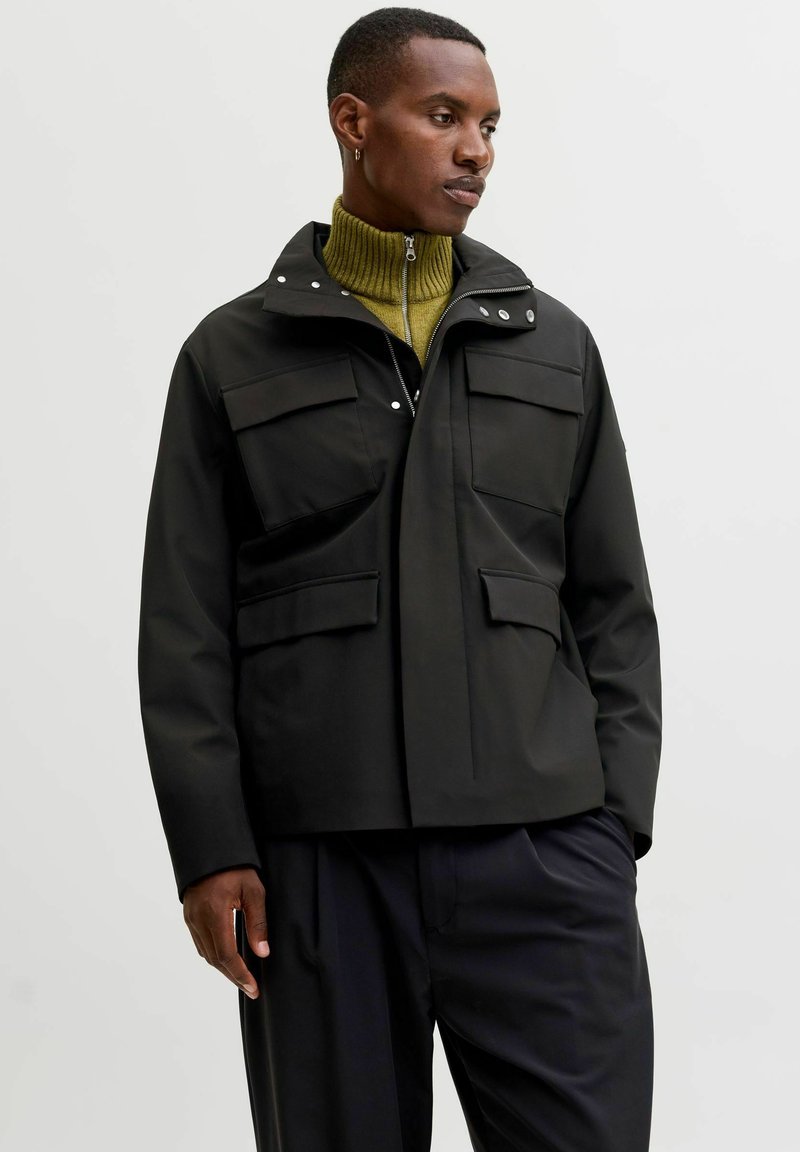 Čierna nepremokavá bunda s vysokým golierom, predným zipsom, patentkami a dvoma veľkými náprsníkovými vreckami. Nosí sa cez zelený rebrovaný rolák.