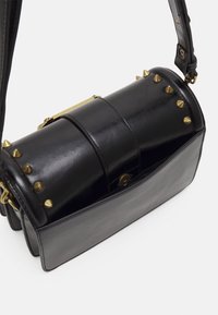 Svart cylindrisk läderhandväska med gyllene koniska nitar längs den övre kanten och justerbar axelrem på vit bakgrund.