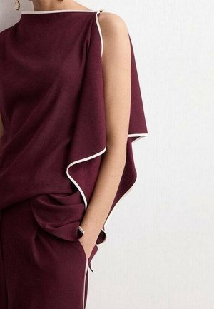 Vrouw draagt een mouwloos kastanjebruin outfit met witte bies, één hand in de broekzak, staand tegen een effen witte muur.