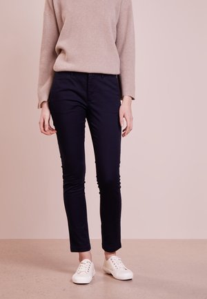 Persoon die een beige geribbelde trui, donkerblauwe smalle broek en witte vetersneakers draagt, staand tegen een effen lichtroze achtergrond.