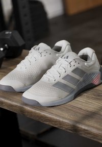 Чифт светлосиви атлетични обувки Adidas с тъмносиви ивици, поставени на дървена повърхност до черна гиричка.