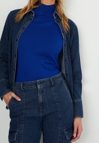 Mörkblå denimskjorta med knappar över en ljusblå ribbad polo, kombinerad med högmidjade denimbyxor med cargofickor.