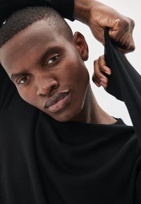 Melnā adītā džemperis ar brīvu, karājošu kakla izgriezumu. Audums izskatās mīksts ar gludu tekstūru. Modelis pozē ar paceltu vienu roku, pielāgojot piedurkni.