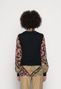 Czarna wełniana kamizelka nałożona na wzorzystą bluzkę z długim rękawem, która ma kręte wzory w kolorze pomarańczowym, zielonym i niebieskim. Gładka tekstura, luźny krój.