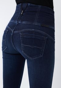 Jeans de tiro alto de color azul oscuro con cierre de cremallera, dos bolsillos traseros y costuras en contraste en un diseño ajustado.