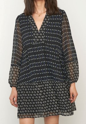 Woman wearing black dress with long sheer sleeves and mixed geometric patterns in white and blue, standing against plain background.