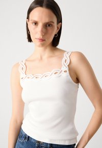 White ribbed tank top featuring decorative lace trim along the neckline and shoulder straps, paired with blue denim jeans.
