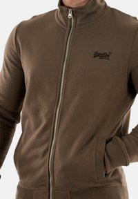 Veste zippée marron en matériau doux, avec un col haut, des poignets côtelés et des poches latérales. Logo discret sur la poitrine.