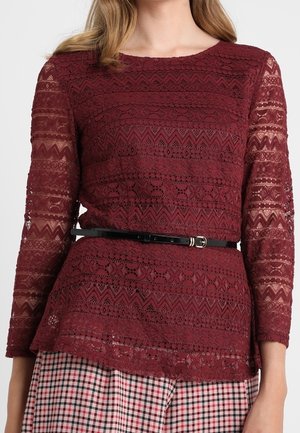 Mujer con un top de encaje burdeos de manga larga, con cinturón negro y pantalones de cuadros en rojo, negro y blanco.