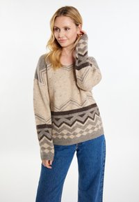 Pulover bej tricotat cu un model geometric în nuanțe de maro și gri. Are guler în V și o croială lejeră, asortat cu blugi din denim albastru.
