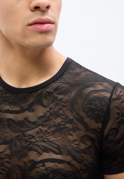 Κοντινό σχέδιο ενός ατόμου που φοράει διάφανο μαύρο τοπ από δαντέλα με λουλουδένια και σπειροειδή σχέδια, εμφανίζοντας το λαιμό, τη γραμμή του σαγονιού και το άνω μέρος του στήθους.