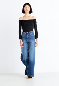 Black off-the-shoulder top, long sleeves; high-waisted, wide-leg blue denim jeans. Model wears black shoes, standing against a white background.