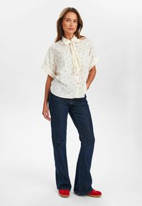 Cream button-up shirt with floral pattern, bow tie detail, short sleeves, paired with dark blue flared jeans and red shoes.
