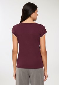 Kobieta z długimi ciemnymi włosami, ubrana w krótką bordową bluzkę z koralikowymi wykończeniami rękawów oraz szare spodnie, pokazana z tyłu na białym tle.