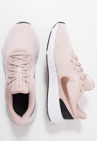 Hellrosa Sportschuhe mit Mesh-Obermaterial, strukturiertem Stoff, weißen Mittelsohlen und schwarzer Außensohle; mit glänzendem roségoldenem Nike-Logo.