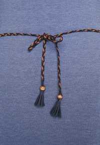 Niebieski teksturowany materiał z zaplecionym czarno-pomarańczowym wiązaniem, ozdobiony dwoma frędzlami i drewnianymi koralikami na końcach.