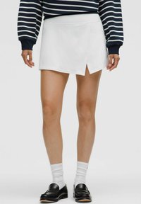 Weißer Skort mit seitlichem Schlitz, der eine glatte Textur und eine taillierte Passform bietet. Kombiniert mit schwarzen Slippern und weißen Socken.