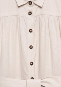 Beige-pitkäliinapaita, jossa on klassinen kaulus, seitsemän tummanruskeaa nappia, rypytetty yksityiskohta rinnan alla ja solmittava vyötärödetail.