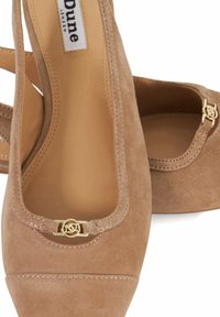 Beige suede pumps featuring a square toe and a gold accent at the strap. Stitching detail along the edges enhances the design.