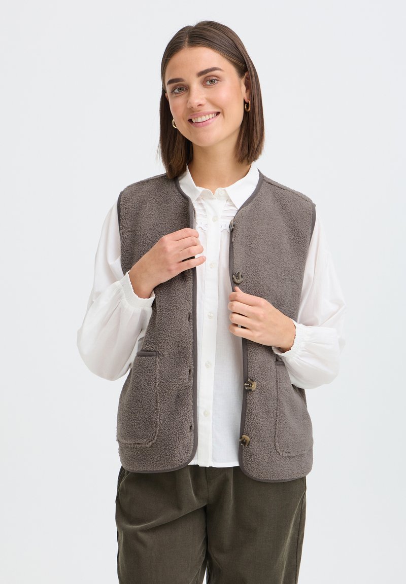 Femme souriante portant un gilet en polaire gris boutonné sur un chemisier blanc et un pantalon sombre, debout devant un fond clair uni.