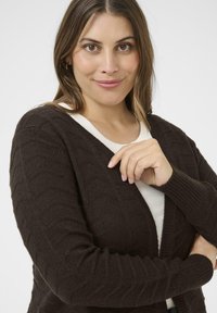 Brauner strukturierter Cardigan mit V-Ausschnitt, langen Ärmeln und komplizierten geometrischen Mustern, über einem weißen Oberteil getragen.