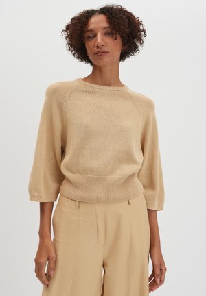 Femme aux cheveux bouclés portant un pull en maille beige à manches trois-quarts et un pantalon beige taille haute, debout devant un fond blanc uni.