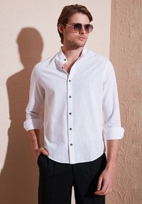 Camisa de lino blanca con cuello alto, cierre de botones frontal, mangas cortas arremangadas y botones oscuros, combinada con pantalones negros.